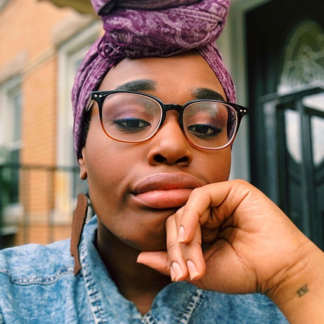 Close up image of Black woman wearing glasses and purple head wrap tied in front knot, and denim shirt. Her hand rests under her chin.